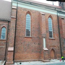 Pro-Builder Paweł Szymański - Mur z czerwonej cegły z gotyckimi oknami i pracownikiem w jaskrawej kamizelce, widoczne ślady renowacji i czyszczenia na dolnej partii ściany.