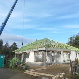 Pro-Builder Paweł Szymański - Wznoszenie domu jednorodzinnego: ściany z betonu komórkowego, drewniana konstrukcja dachu w trakcie montażu, żuraw budowlany w tle, przenośna toaleta na pierwszym planie.