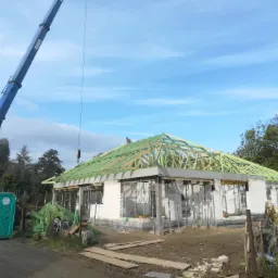 Wznoszenie domu jednorodzinnego: ściany z betonu komórkowego, drewniana konstrukcja dachu w trakcie montażu, żuraw budowlany w tle, przenośna toaleta na pierwszym planie.
