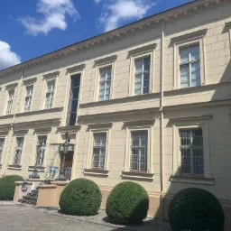 Odnowiona elewacja budynku w stylu klasycystycznym z licznymi oknami z kratami, widoczne kuliste krzewy przed budynkiem i fragment błękitnego nieba z chmurami.