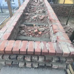 Budowa muru z cegły z widocznymi elementami rusztowania i gruzem wewnątrz konstrukcji, w tle budynki miejskie.