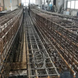 Seryjnie przygotowane szkielety z prętów zbrojeniowych ułożone równolegle w hali produkcyjnej, widoczne elementy drewniane i metalowe, w tle okna i sylwetki pracowników.