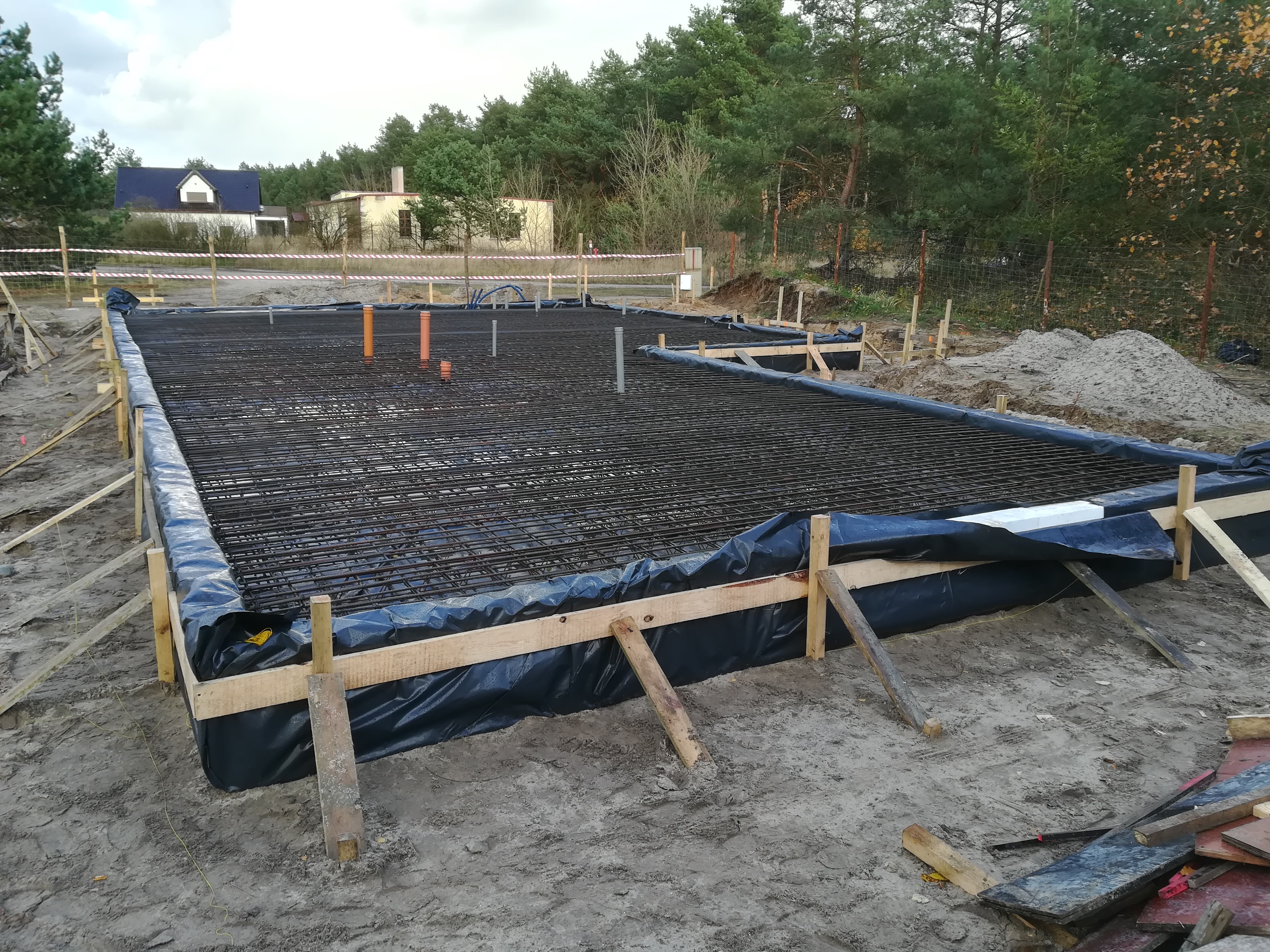 Przygotowanie zbrojenia fundamentu pod budynek, widoczne drewniane szalunki, czarna folia izolacyjna, siatka z prętów zbrojeniowych oraz wystające rury instalacyjne. W tle zabudowa jednorodzinna...