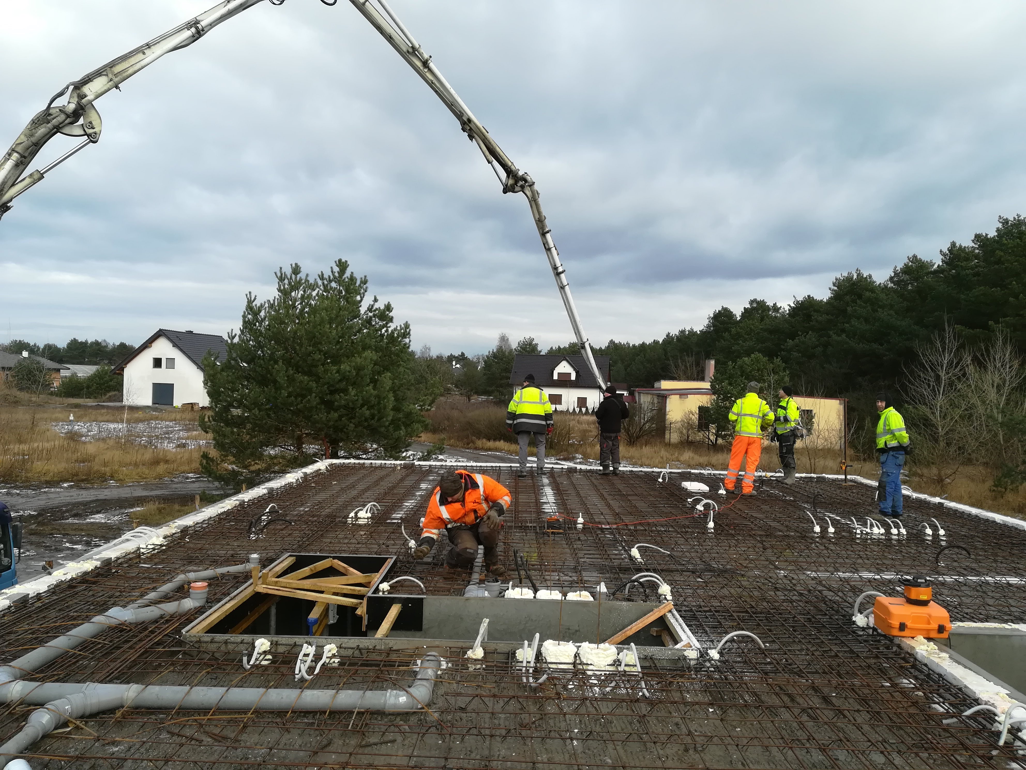 Prace na budowie w Lipnie: Ekipa budowlana w kamizelkach odblaskowych przygotowuje zbrojenie stropu z widocznymi instalacjami, z betonową pompą w tle i domami w oddali.