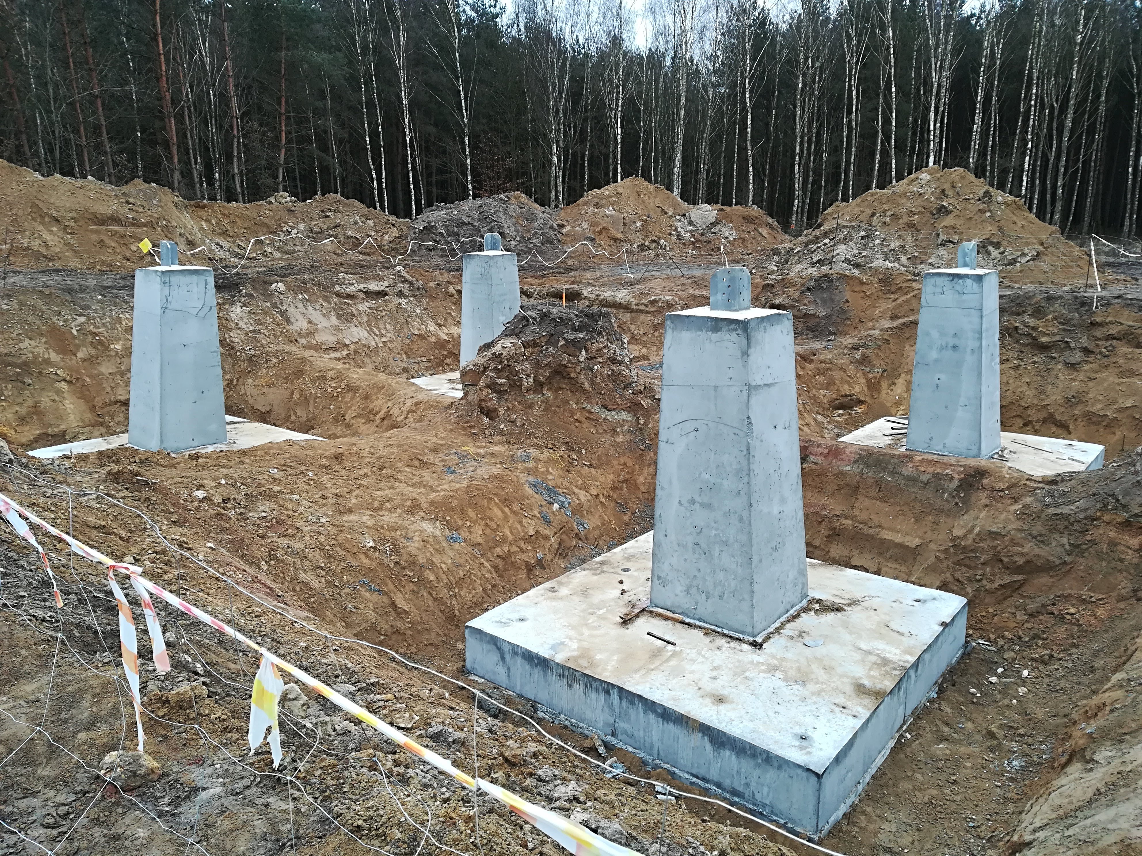 Betonowe słupy fundamentowe na placu budowy, widoczne wykopane rowy i taśmy ostrzegawcze, w tle las.