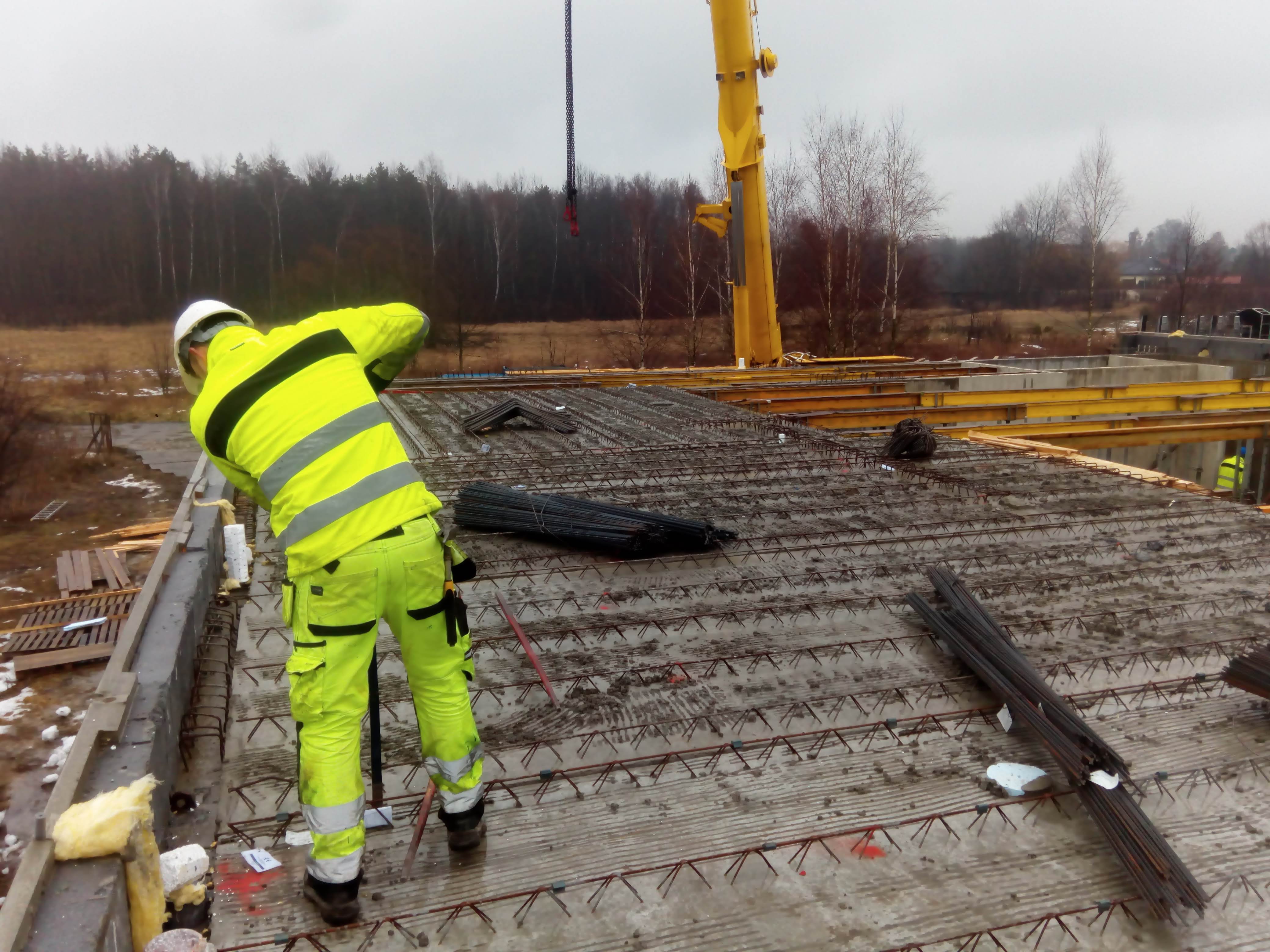 Zbrojarz w jaskrawej odzieży roboczej pochyla się nad zbrojeniem płyty fundamentowej, w tle widoczny żuraw budowlany i drzewa.