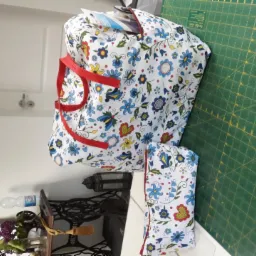 Torba zakupowa i kosmetyczka uszyte z białej tkaniny w kwiaty, z czerwonymi uchwytami i lamówką, leżą na zielonej macie do cięcia, w tle maszyna do szycia.