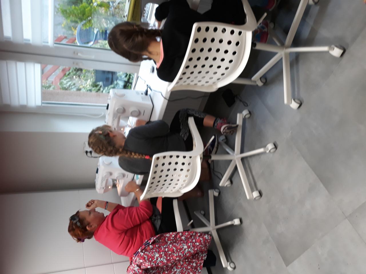 Trzy osoby, w tym instruktor, siedzą przy maszynach do szycia podczas zajęć z szycia w jasnym pomieszczeniu, z widokiem na ogród przez okno.