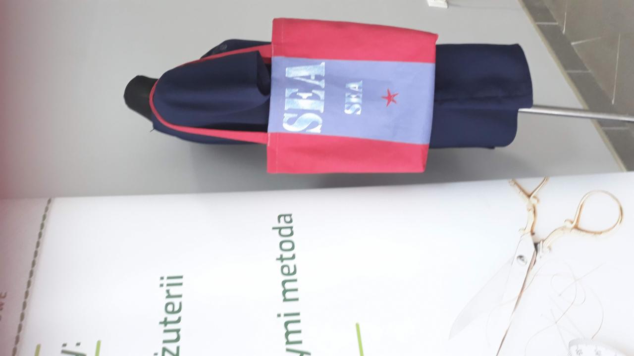 Manekin w granatowym płaszczu z czerwoną lamówką, na którym zawieszona jest torba z napisem 'SEA' i czerwoną gwiazdą, obok leżą złote nożyczki krawieckie i miarka.