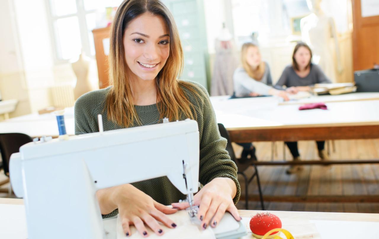 Uśmiechnięta młoda kobieta szyje na białej maszynie do szycia w jasnej pracowni, w tle dwie osoby przy stole i manekin.