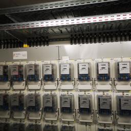 Volt Electro - Rząd liczników elektrycznych zamontowanych w szafie rozdzielczej z widocznymi przewodami i elementami mocującymi na metalowej konstrukcji nośnej.