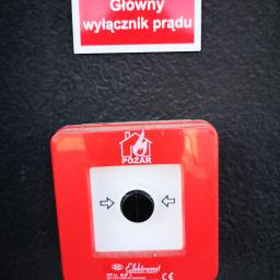 Volt Electro - Czerwony przycisk alarmowy 'Pożar' z czarnym okrągłym włącznikiem, umieszczony pod tabliczką 'Główny wyłącznik prądu' na ciemnej, teksturowanej ścianie.
