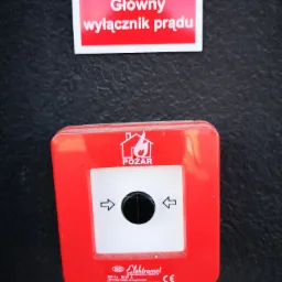 Czerwony przycisk alarmowy 'Pożar' z czarnym okrągłym włącznikiem, umieszczony pod tabliczką 'Główny wyłącznik prądu' na ciemnej, teksturowanej ścianie.