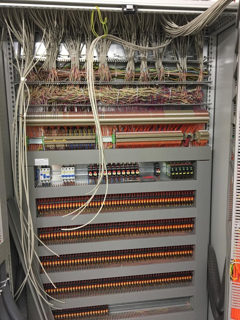 Otwarta szafa sterownicza z bogatym okablowaniem, widoczne liczne przewody, złączki i elementy zabezpieczające, ułożone w rzędach, niektóre kable luźno zwisają.