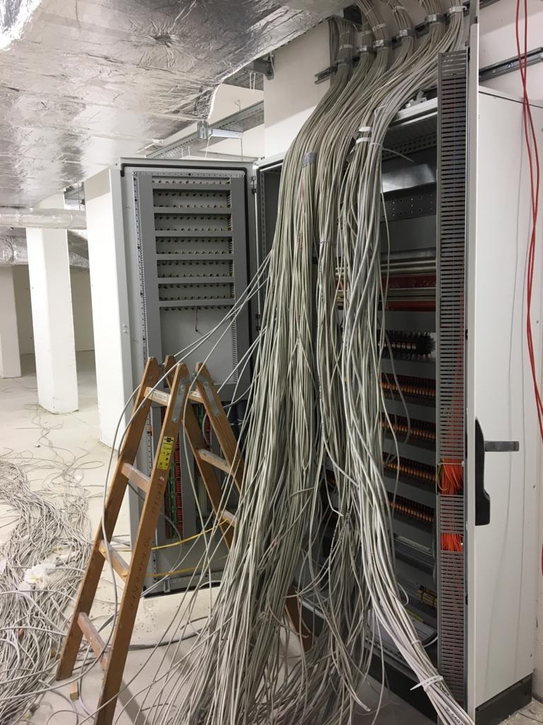 Otwarta szafa sterownicza z licznymi kablami wchodzącymi od góry, drabina drewniana oparta o szafę, sufit z widoczną instalacją, stan surowy.