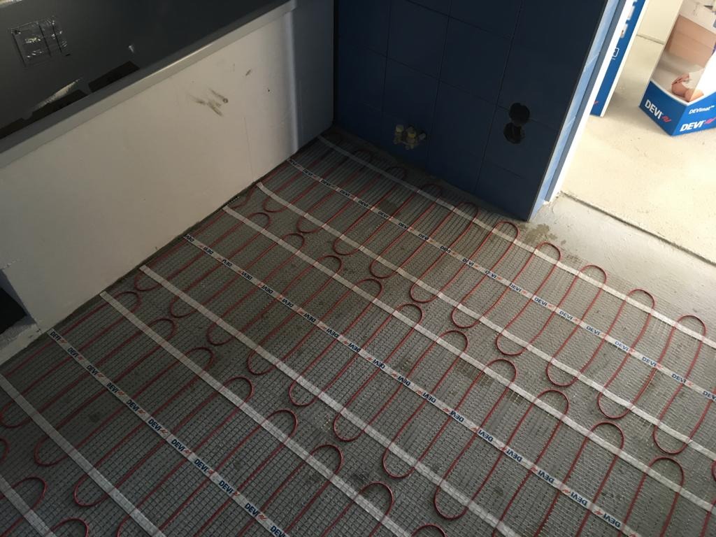 Montaż ogrzewania podłogowego elektrycznego marki Devi w łazience z niebieskimi płytkami, widoczne kable grzewcze na siatce i fragment wanny.