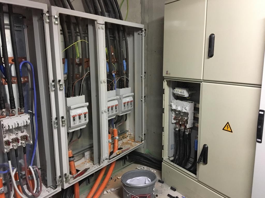 Otwarta szafa elektryczna z widocznymi zabezpieczeniami, przewodami i elementami instalacji, obok zamknięta szafa z symbolem ostrzegawczym, na podłodze resztki prac instalacyjnych i wiadro.