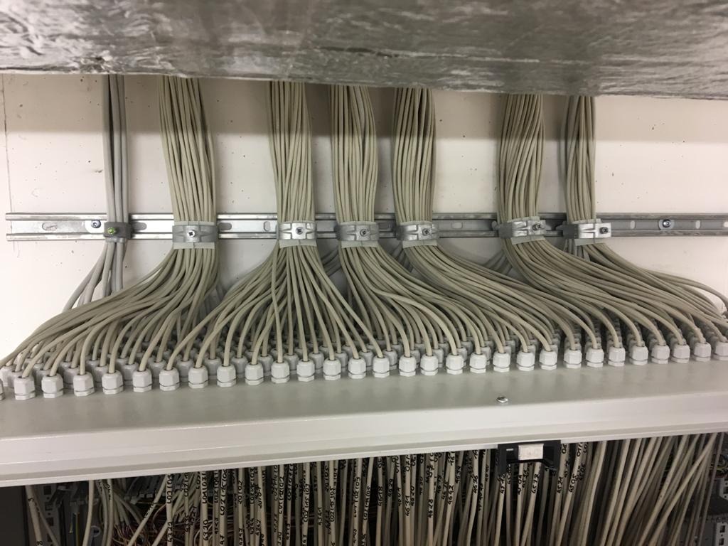 Równolegle ułożone wiązki kabli elektrycznych, zabezpieczone uchwytami do szyny montażowej, z widocznymi numerami identyfikacyjnymi na dolnej sekcji.