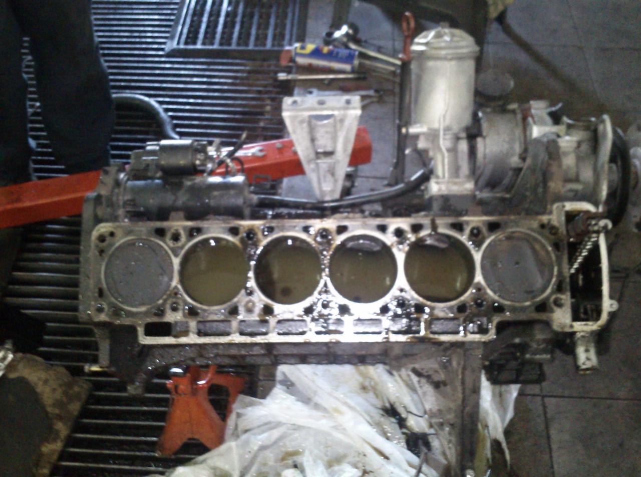 Blok silnika samochodowego z widocznymi cylindrami wypełnionymi płynem, umieszczony na czerwonym podnośniku warsztatowym, z widocznymi narzędziami w tle.