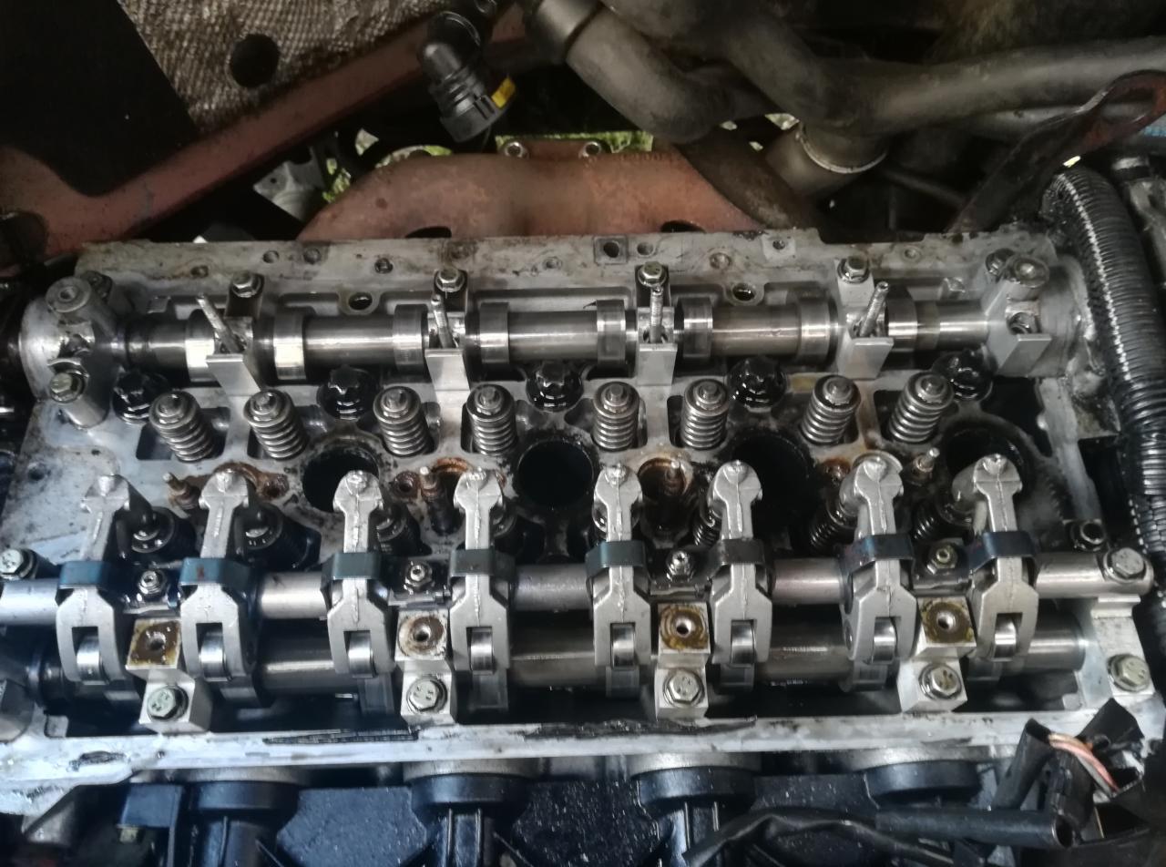 Widok z bliska na otwarty silnik samochodowy z widocznymi wałkami rozrządu, sprężynami zaworowymi i dźwigienkami, pokryty olejem, ukazujący etap naprawy lub konserwacji.