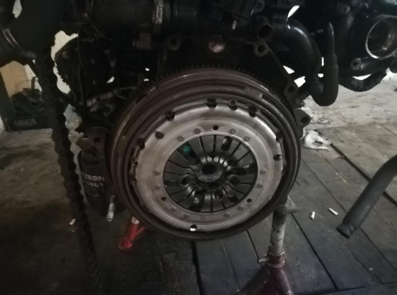 Zdjęcie sprzęgła samochodowego zamontowanego na silniku, widoczne ślady użytkowania i zabrudzenia, silnik podparty na czerwonym stojaku warsztatowym, w tle podłoga z desek i części samochodowe.