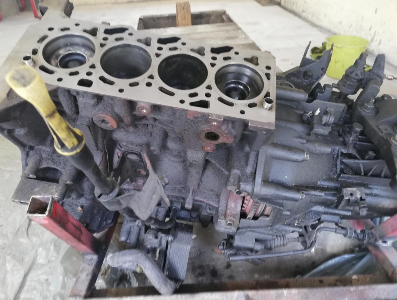 Brudny, częściowo zdemontowany blok silnika na metalowym stojaku, z widocznymi otworami cylindrów i żółtym bagnetem oleju.