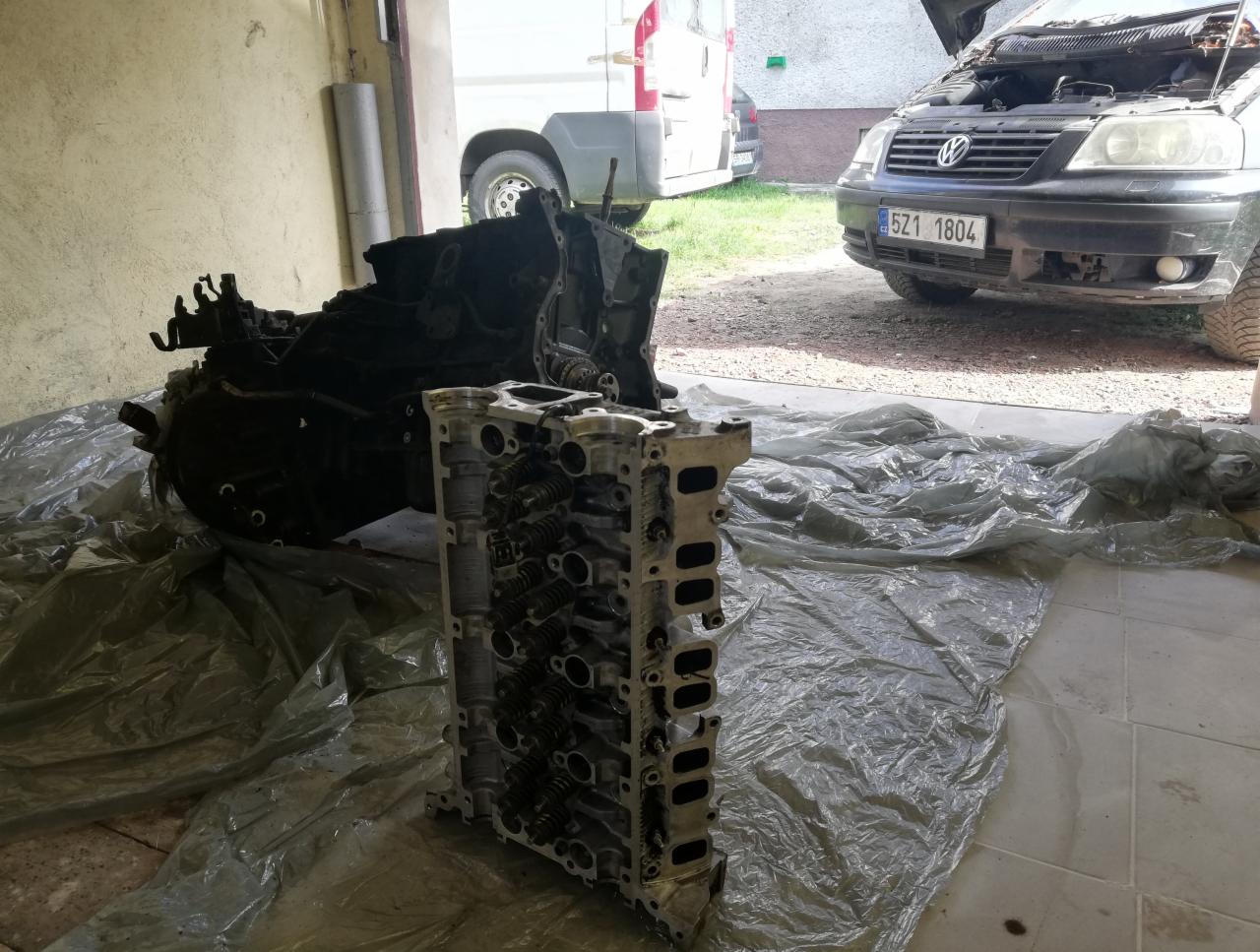 Wyjęty silnik samochodowy i głowica cylindrów leżące na folii w warsztacie, z otwartą maską samochodu widoczną w tle.