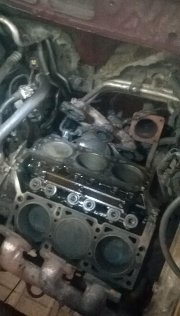 Widok z góry na zdemontowany blok silnika samochodowego, z widocznymi cylindrami i elementami osprzętu, w otoczeniu przewodów i części w komorze silnika.