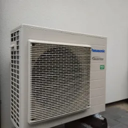 Zewnętrzna jednostka pompy ciepła Panasonic Aquarea Inverter R32 zamontowana na szarym cokole przy jasnej ścianie budynku.