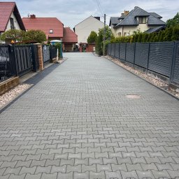 KGBUD Grzegorz Kunicki - Długa, szara droga z kostki brukowej między ogrodzeniami posesji. Kostka ułożona w regularny wzór, widoczne domy w tle i pochmurne niebo. Estetyczne wykończenie.