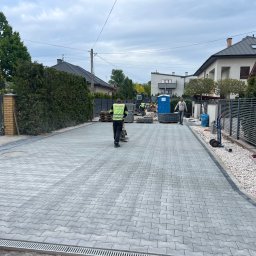 KGBUD Grzegorz Kunicki - Układanie szarej kostki brukowej na posesji. Widoczny pracownik zagęszczający powierzchnię oraz ekipa brukarska w tle. Nowoczesna zabudowa jednorodzinna.