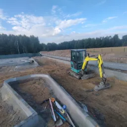 Narożne obrzeża betonowe w gruncie, minikoparka, narzędzia. Przygotowanie terenu pod budowę drogi lub chodnika na tle zieleni i błękitnego nieba.