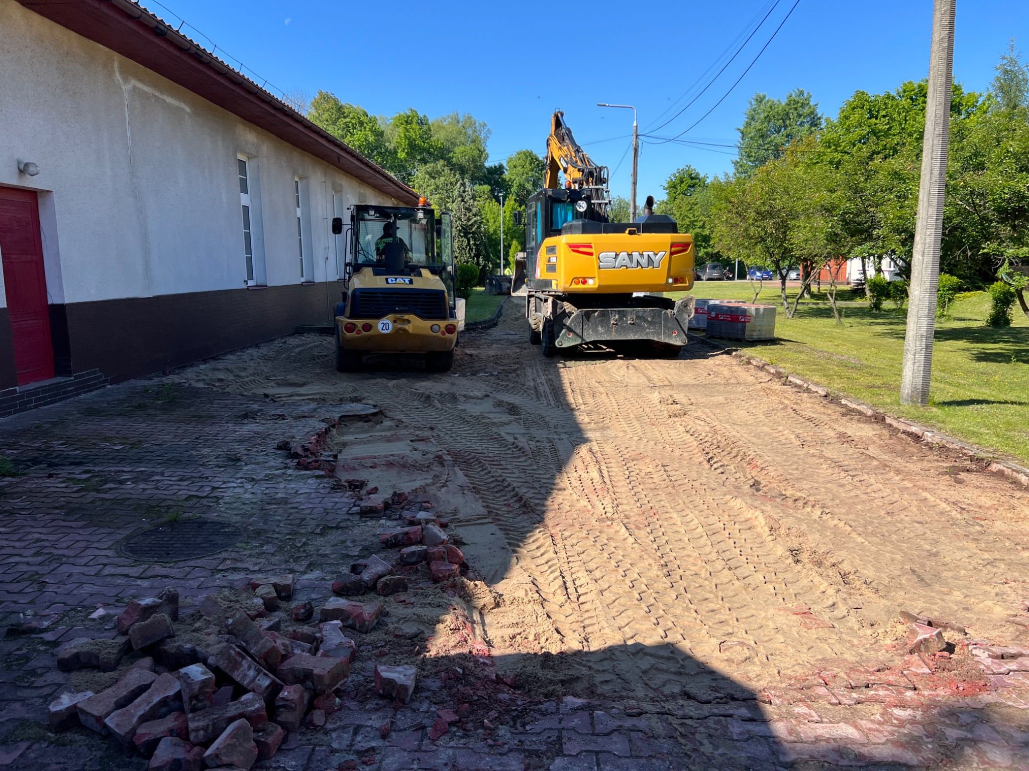 Żółty walec CAT i koparka SANY na placu budowy. Częściowo usunięta kostka brukowa, przygotowanie podłoża. Widoczne ślady opon na piasku. Budynek w tle.