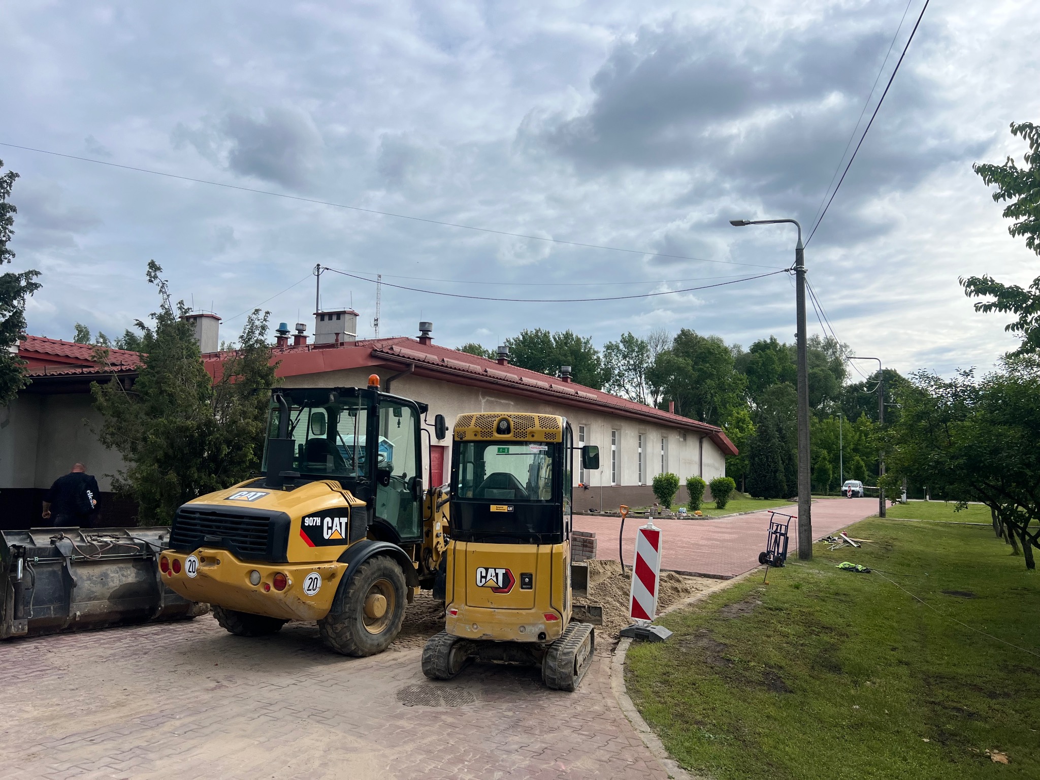 Żółty ładowacz czołowy CAT 907H i minikoparka CAT na tle budynku z czerwoną dachówką i ułożonej kostki brukowej. Prace ziemne w toku.
