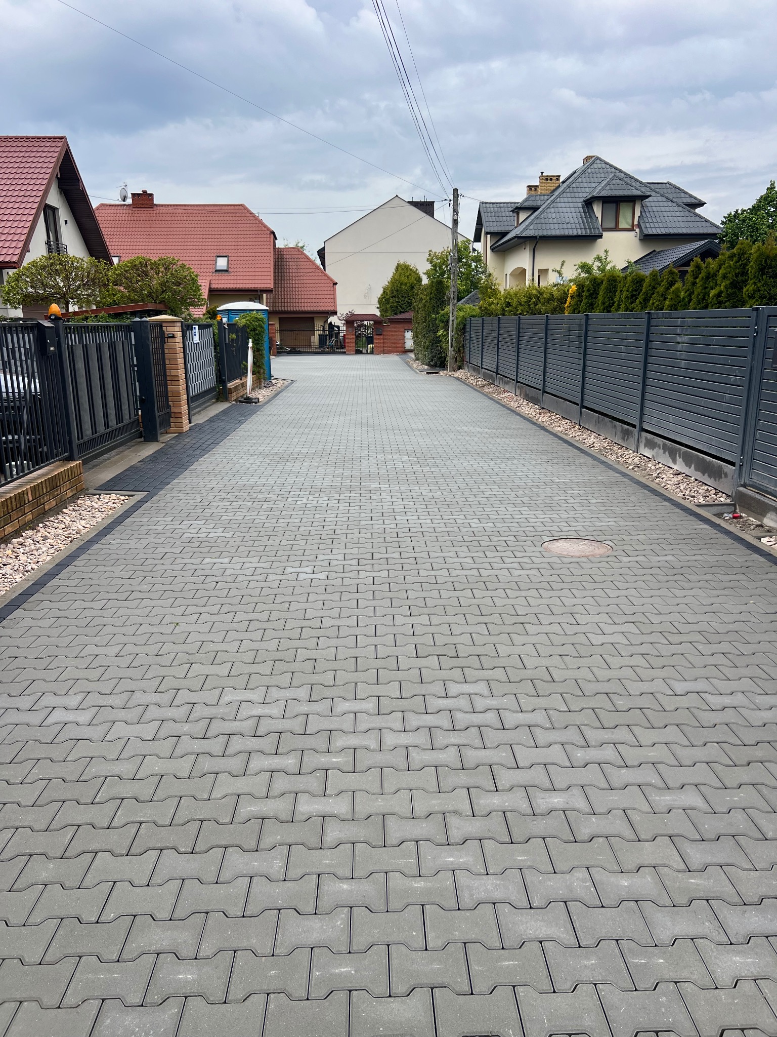 Długa, szara droga z kostki brukowej między ogrodzeniami posesji. Kostka ułożona w regularny wzór, widoczne domy w tle i pochmurne niebo. Estetyczne wykończenie.