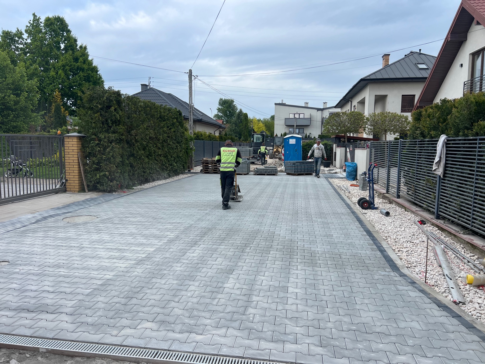 Układanie szarej kostki brukowej na posesji. Widoczny pracownik zagęszczający powierzchnię oraz ekipa brukarska w tle. Nowoczesna zabudowa jednorodzinna.