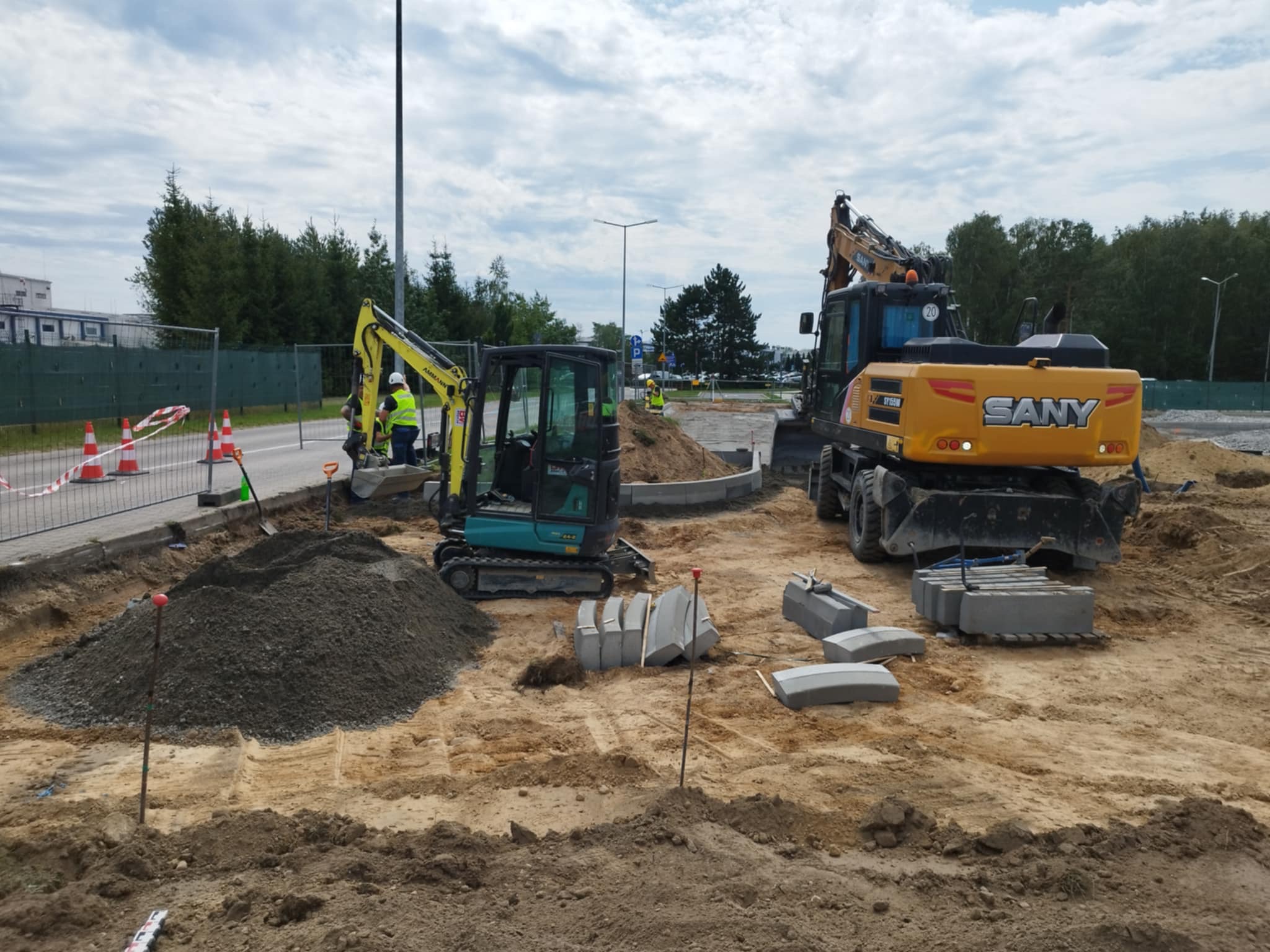 Dwa żółte i turkusowe minikoparki na placu budowy z przygotowanymi elementami betonowymi. W tle widoczne drzewa i błękitne niebo z chmurami.