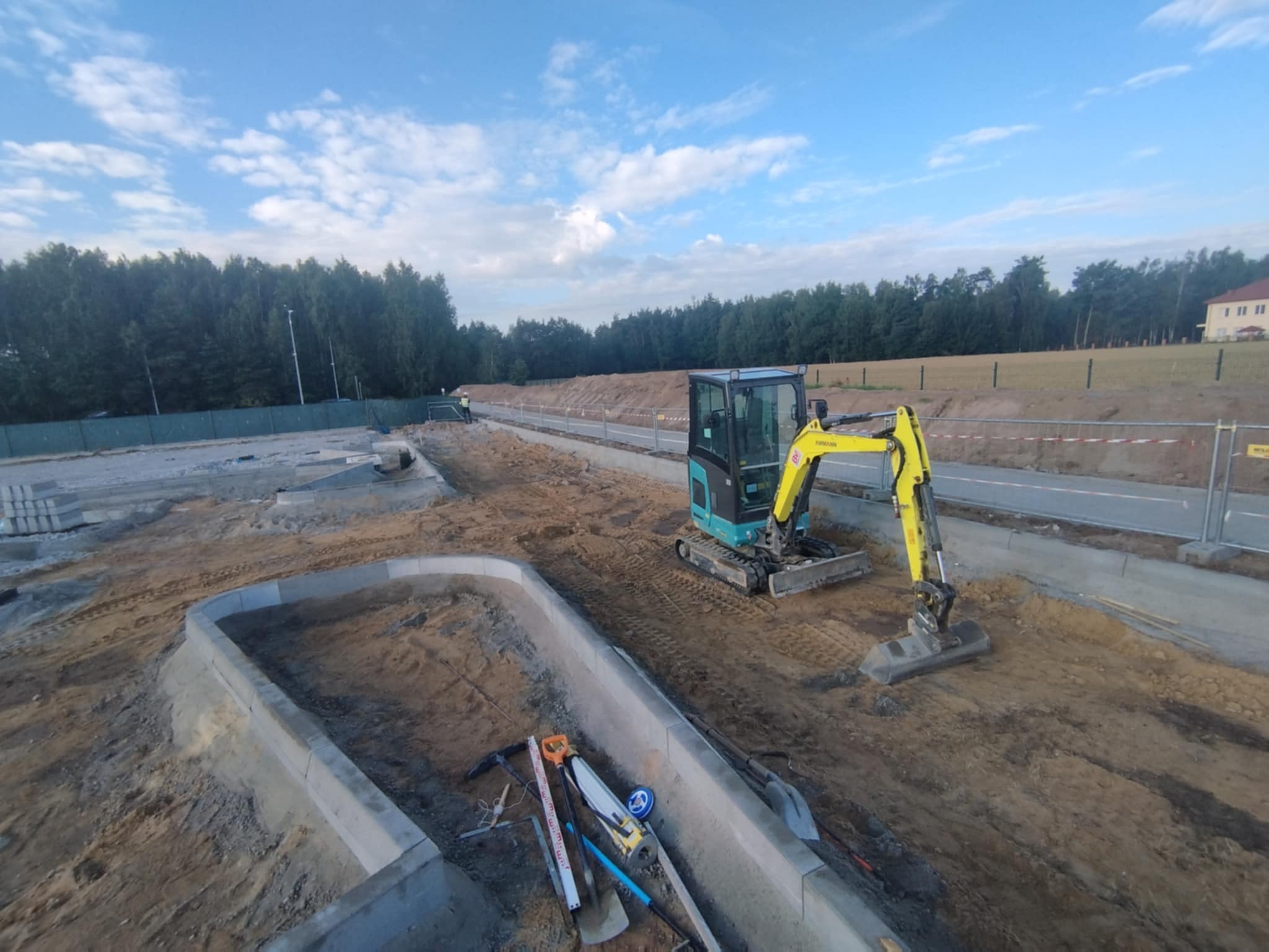 Narożne obrzeża betonowe w gruncie, minikoparka, narzędzia. Przygotowanie terenu pod budowę drogi lub chodnika na tle zieleni i błękitnego nieba.