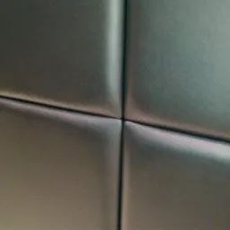 Fragment pikowanej, ciemnoszarej skóry, prawdopodobnie element tapicerki samochodowej lub meblowej, widoczne szwy krzyżujące się pod kątem prostym.