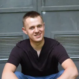 Uśmiechnięty młody mężczyzna w czarnej koszulce i niebieskich spodniach siedzi na schodach na tle szarej, metalowej ściany z kratką wentylacyjną i zamkiem na górze.