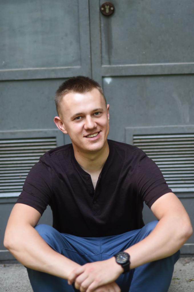 Uśmiechnięty młody mężczyzna w czarnej koszulce i niebieskich spodniach siedzi na schodach na tle szarej, metalowej ściany z kratką wentylacyjną i zamkiem na górze.