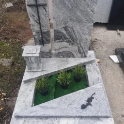 Jasnoszary nagrobek z granitu z czarnym krzyżem i różą, trzy doniczki z zielonymi roślinami na sztucznej trawie.