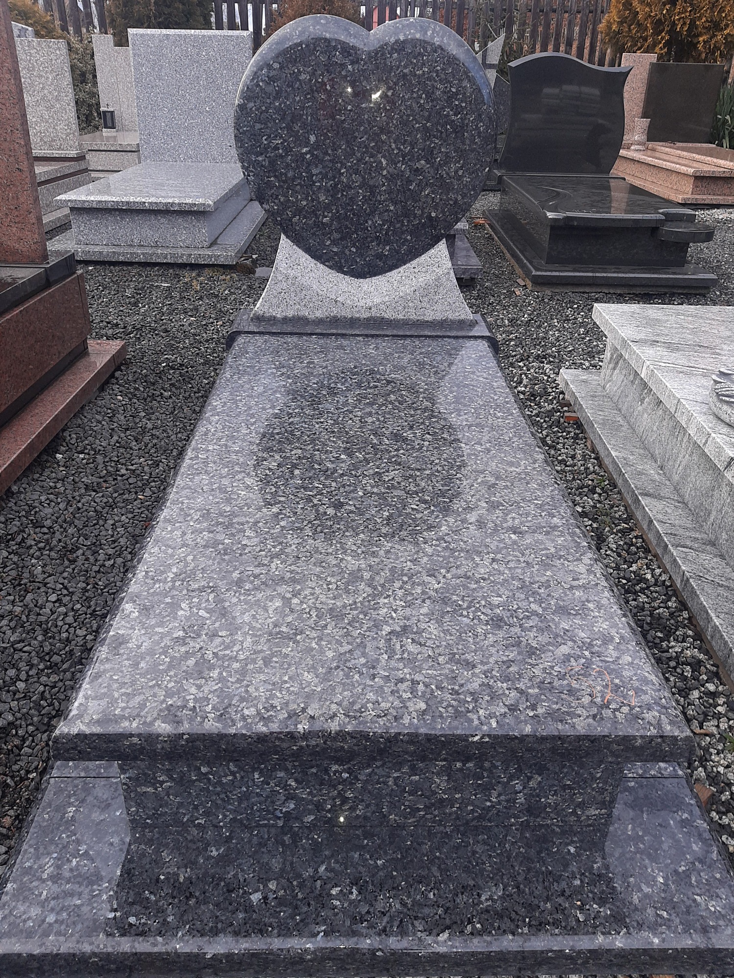 Polerowany, ciemnoszary pomnik z sercem na szczycie, otoczony żwirem i innymi nagrobkami na zewnątrz.