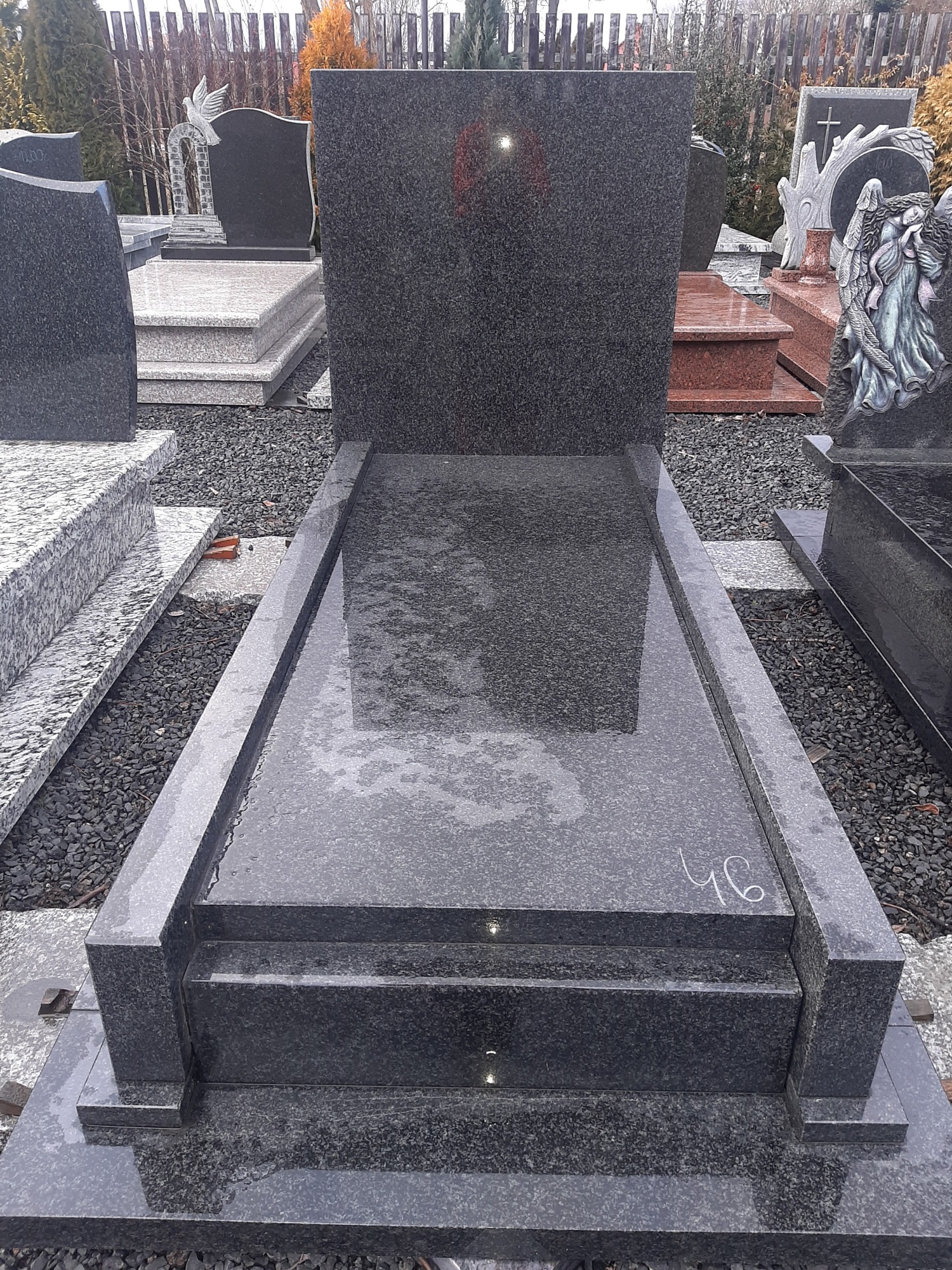 Nowoczesny, ciemnoszary nagrobek z granitu z wyraźnym odbiciem otoczenia na mokrej powierzchni, ustawiony na żwirowym podłożu wśród innych nagrobków na ekspozycji.