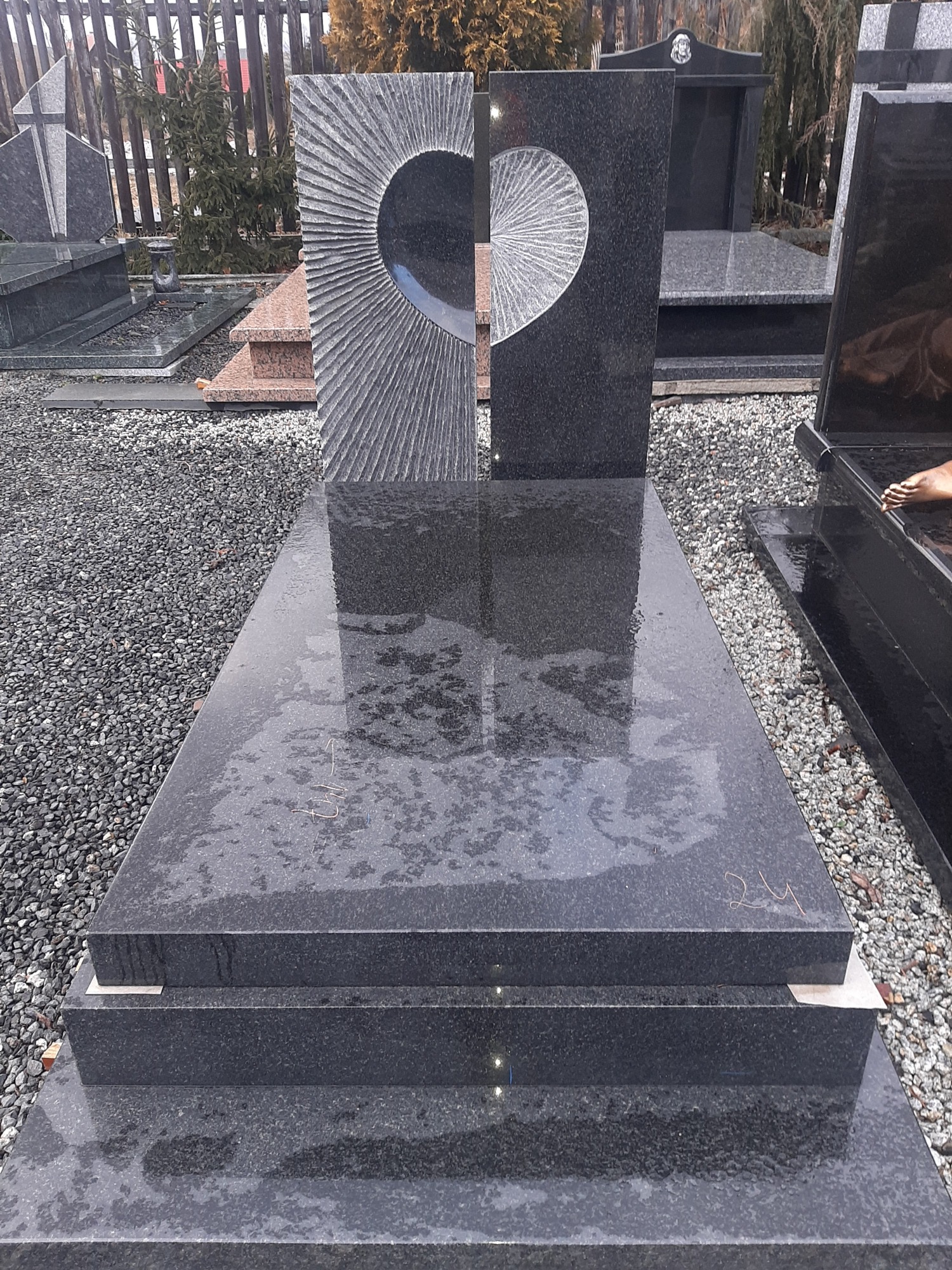 Nowoczesny, czarny nagrobek z granitu z wyciętym motywem połowy serca z promienistym wzorem, widoczny na tle innych nagrobków i żwirowego podłoża.