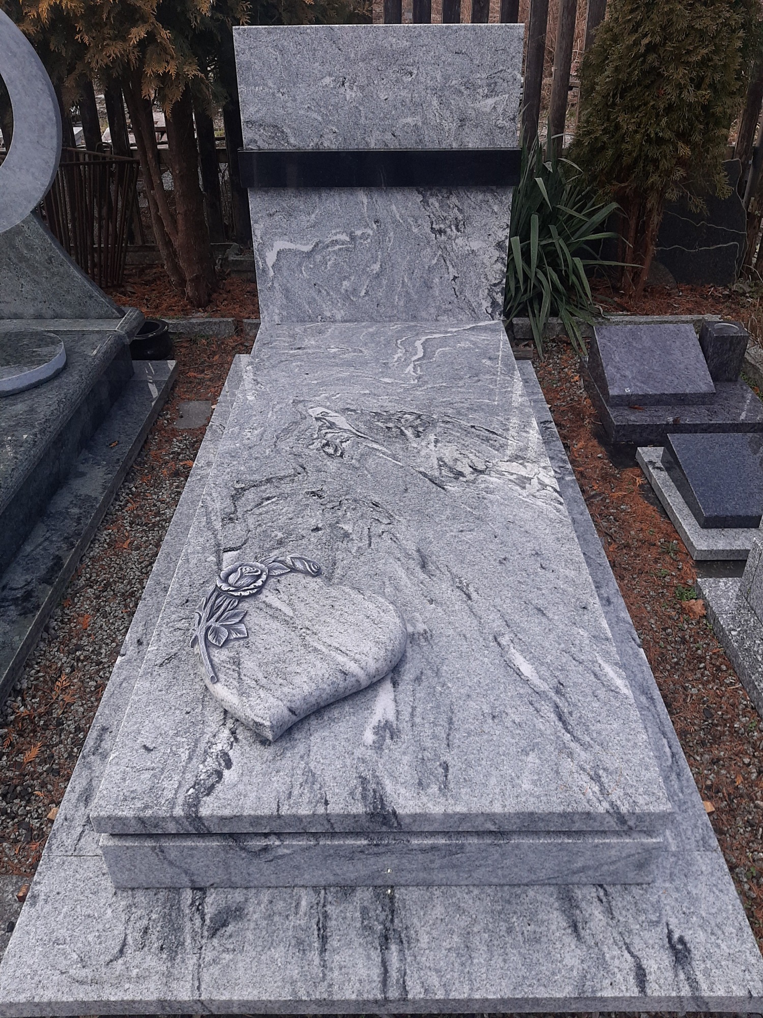 Jasnoszary nagrobek z granitu z motywem serca i róży, na tle cmentarza.