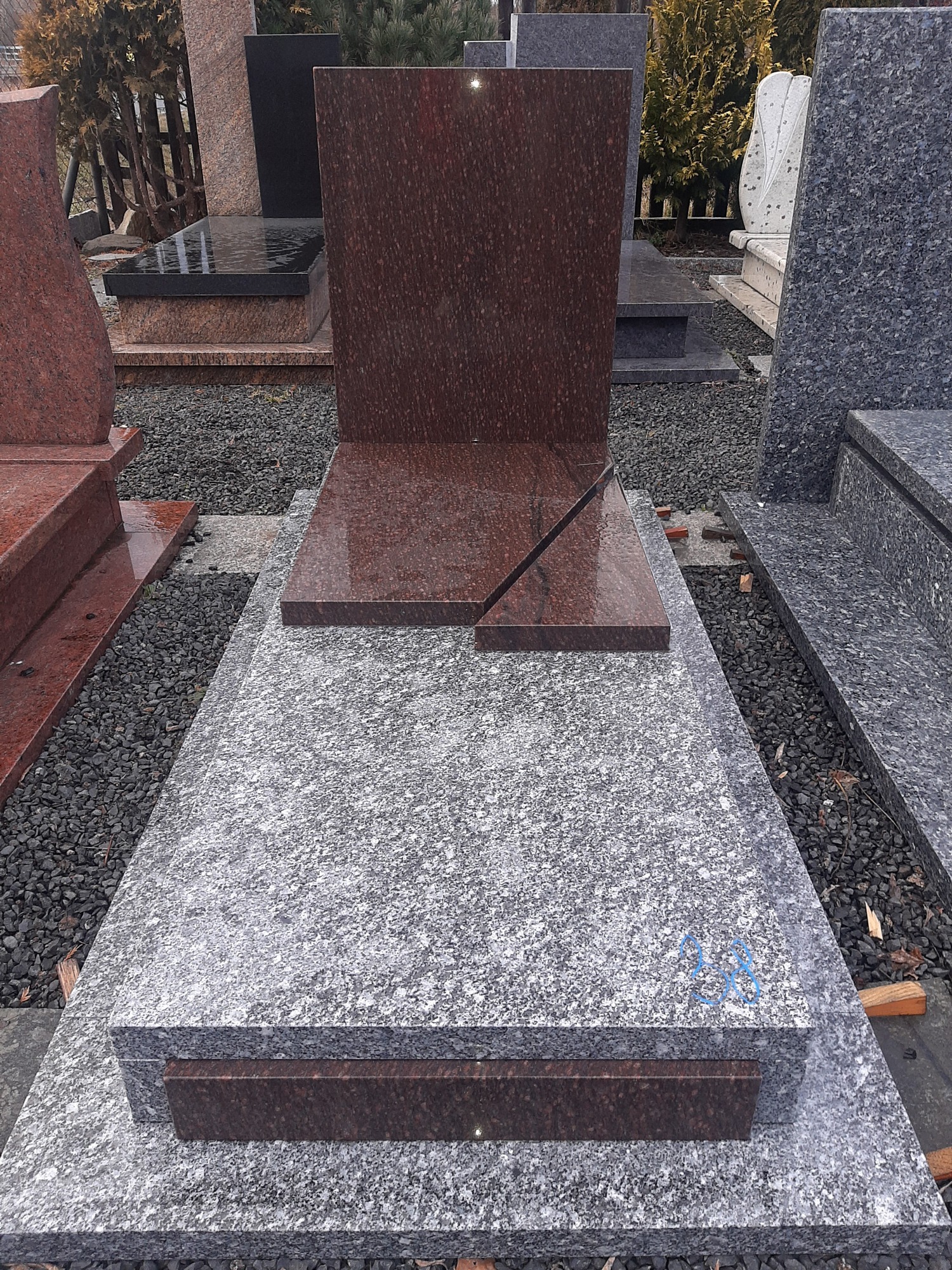 Szary, granitowy pomnik z elementami z czerwonego granitu, z ukośnym przecięciem na płycie nagrobnej. W tle inne nagrobki i drzewa.