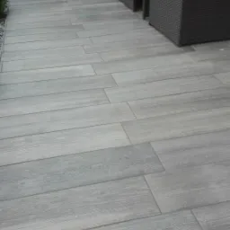 Szare płytki imitujące drewno ułożone na tarasie, z widocznymi meblami ogrodowymi z technorattanu w tle i zielenią.