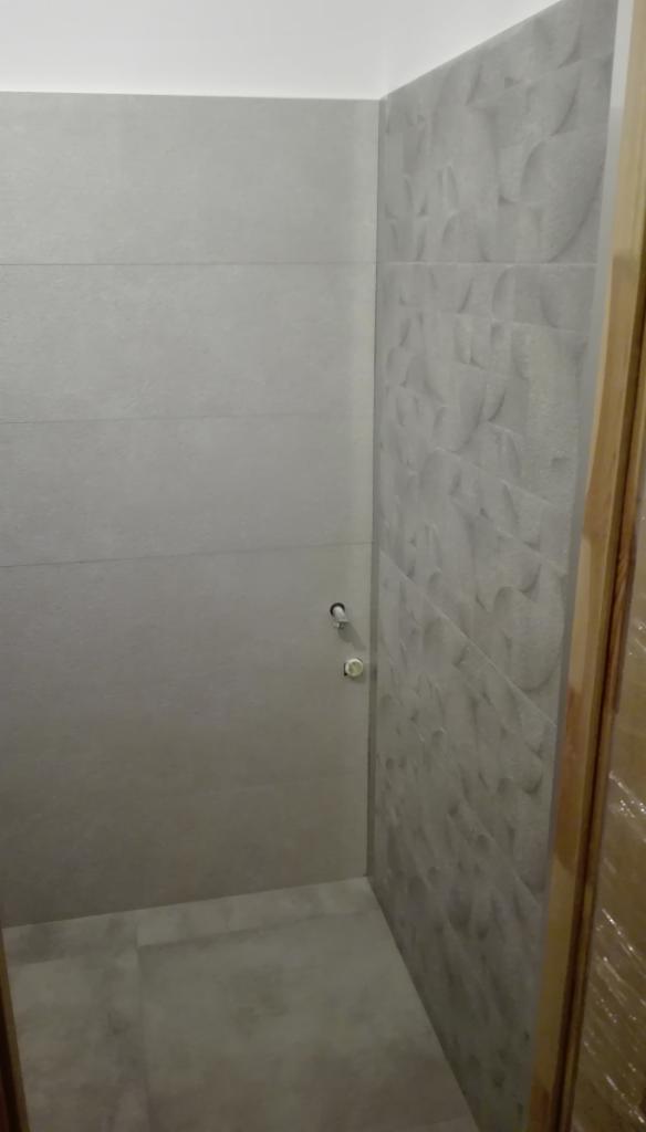 Wykończona łazienka z szarymi, prostokątnymi płytkami na ścianie po lewej i dekoracyjnymi, szarymi płytkami z wzorem na ścianie po prawej, widoczne dwa zawory na ścianie.