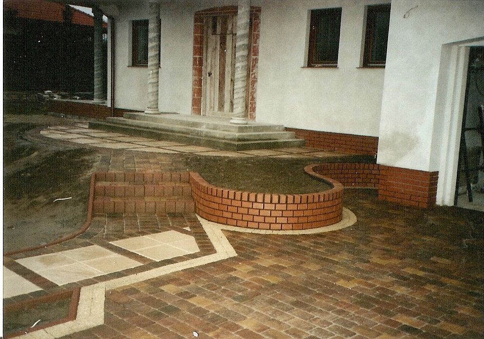 Ułożona kostka brukowa w Muchnowie13, tworząca wzór z beżowych i ceglanych elementów. Widoczne schody z cegły prowadzące do domu z kolumnami.